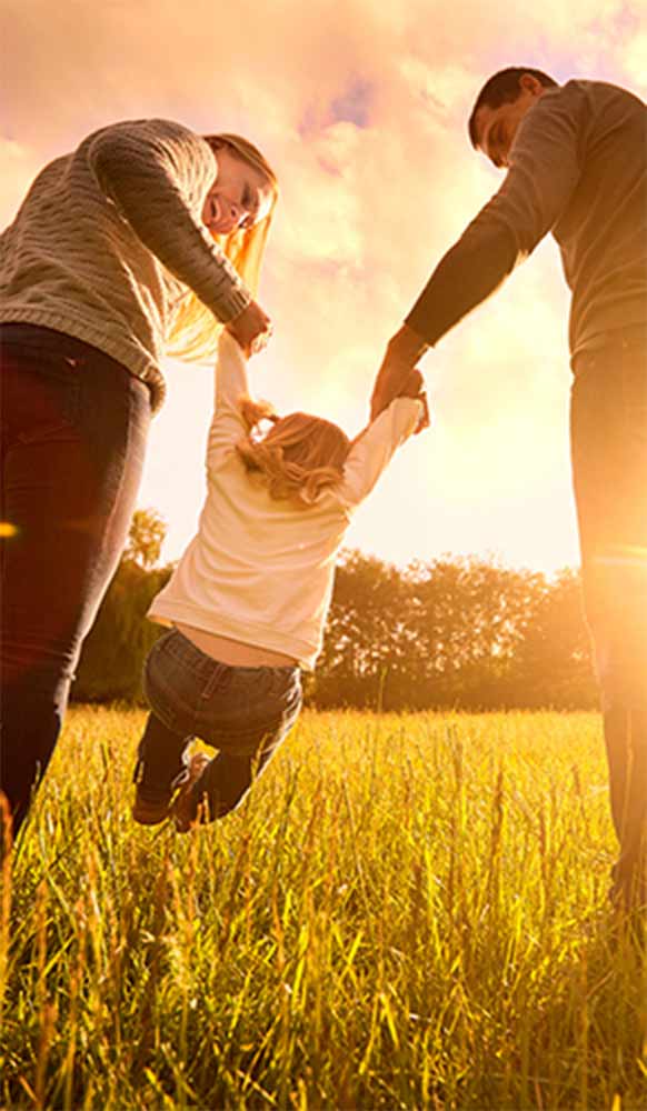 A small family swinging a small child in a grassy field during the Golden Hour